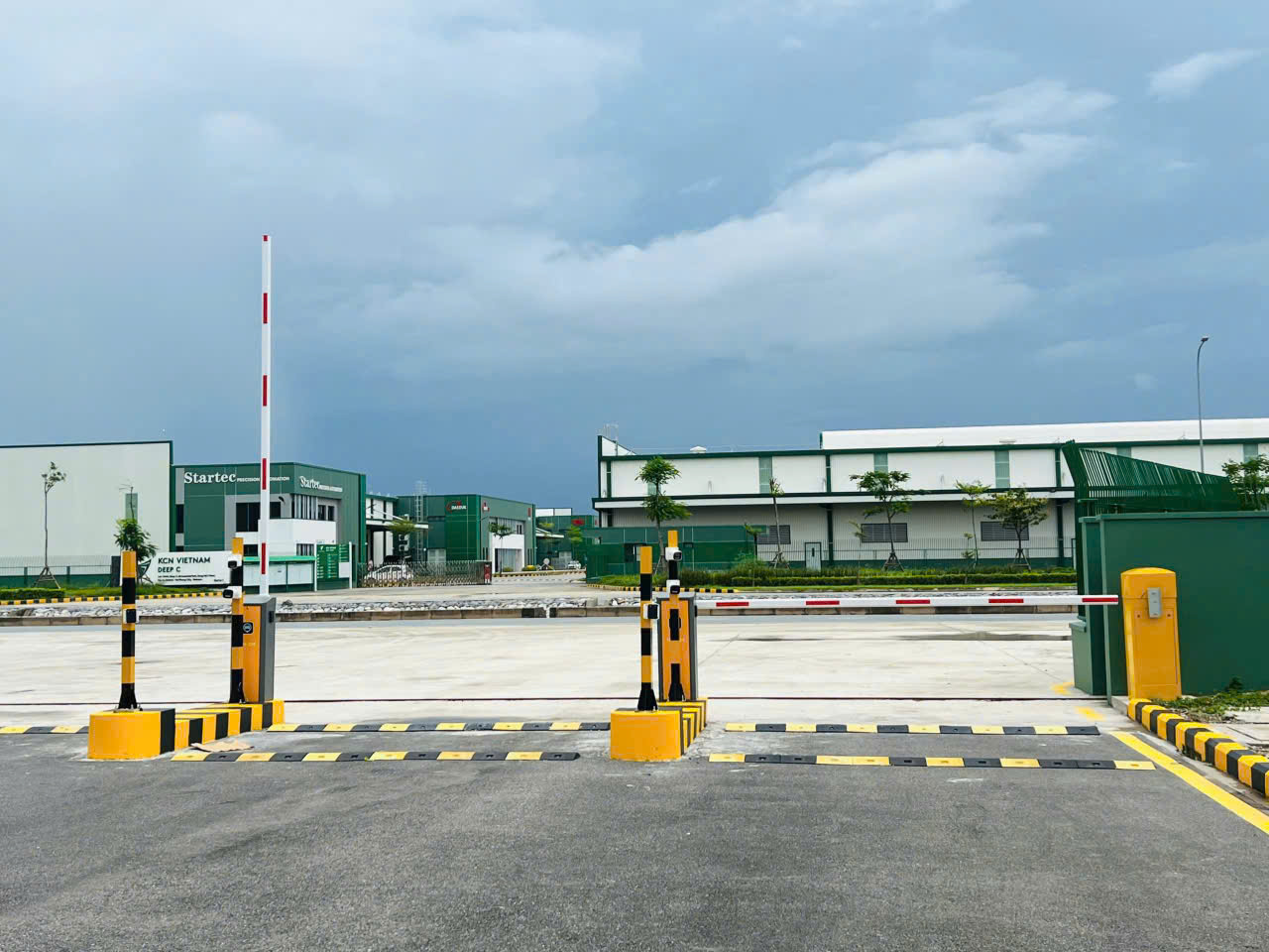 Cổng barie tự động màu vàng đen tại lối vào khu công nghiệp với nhà xưởng phía sau
